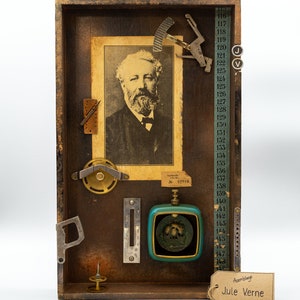 Könnte beinhalten: Eine Holzkiste mit einer Sammlung von Vintage-Objekten, darunter ein Porträt von Jules Verne, ein Kompass, ein Lineal und eine kleine Uhr. Die Kiste ist mit "Assemblage Jule Verne" beschriftet.