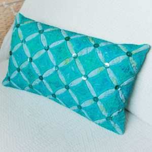 以下が含まれることがあります： ターコイズとブルーのバティック柄の長方形の装飾用枕。枕は円形の幾何学模様で、小さくて丸い虹色のボタンで装飾されています。枕は白い表面に置かれています。