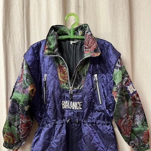 Peut inclure: Une veste matelassée violette avec un imprimé floral sur les manches et un logo "BALANCE" blanc sur le devant. La veste a une taille à cordon et une fermeture éclair.