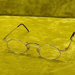 May include: Gold-rimmed eyeglasses with oval lenses. The frames have a delicate design along the edges of the lenses and black accents on the temples. The glasses are set against a green background.