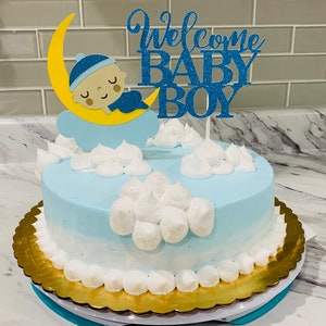 Puede incluir: Un pastel de baby shower azul y blanco con un adorno azul y amarillo de 'Bienvenido Bebé Niño'. El pastel está decorado con nubes de glaseado blanco y un bebé durmiendo en una luna creciente amarilla.