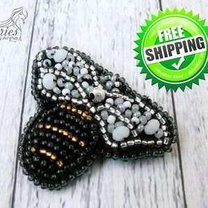 Pode incluir: Um broche em forma de abelha feito à mão com trabalho intrincado de missangas. As asas são compostas por missangas brancas translúcidas e prateadas, enquanto o corpo exibe filas alternadas de missangas cinzento-escuras e dourado metálico.
