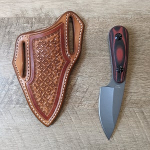 Puede incluir: Una funda de cuchillo de cuero marrón con un diseño estampado y un cuchillo con mango negro y rojo con una hoja gris.