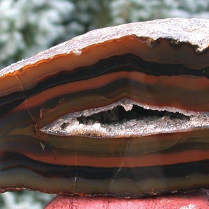 May include: A large, brown agate geode with a natural opening revealing a crystal-lined interior. The agate has a layered, banded pattern in shades of brown, orange, and white.