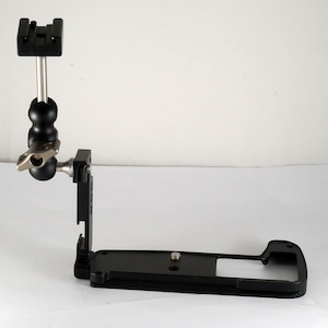 May include: Black camera cage with a vertical arm and a ball head. The arm is attached to the cage with a screw. The ball head has a small knob for adjusting the angle.
