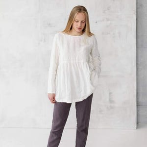 May include: A white linen blouse with long sleeves and a gathered waist. The blouse is worn with gray pants and brown leather sandals.