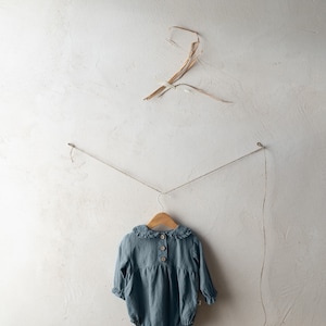 May include: A light blue baby onesie with a ruffled collar and button closure hangs on a wooden hanger against a white wall.
