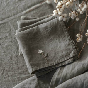May include: A set of four grey linen napkins with a delicate white flower embroidered on one of the napkins.