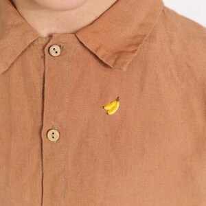 May include: A brown linen shirt with a small embroidered banana design on the front. The shirt has a button-up collar and two buttons on the front.