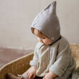Puede incluir: Un bebé con un gorro de lino gris está sentado en una cesta de mimbre. El bebé lleva un suéter gris claro y un vestido blanco. El bebé está mirando hacia abajo a la cesta.