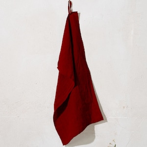 Puede incluir: Un paño de cocina de lino rojo oscuro colgado de un gancho contra una pared blanca.