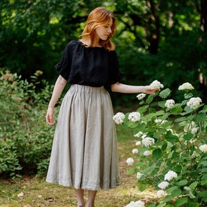 Puede incluir: Una mujer con una blusa de lino negra y una falda de lino marrón claro está de pie en un jardín. Está extendiendo la mano para tocar una flor blanca.