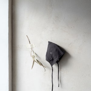 Puede incluir: Un gorro de bebé de lino gris oscuro con una estrella blanca bordada colgando de una pared blanca texturizada.