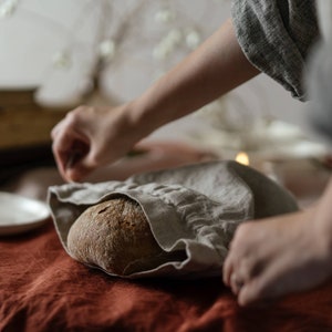 Könnte beinhalten: Eine Person verpackt ein Sauerteigbrot in ein Leinen-Tuch. Das Brot ist braun und knusprig, und das Tuch ist hellbeige. Die Hände der Person sind sichtbar, und der Hintergrund ist verschwommen.