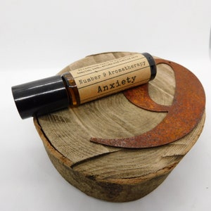 May include: A brown glass roller bottle with a black cap and a label that reads "Number 9 Aromatherapy Anxiety". The bottle is sitting on a wooden surface with a rusty metal crescent moon shape.