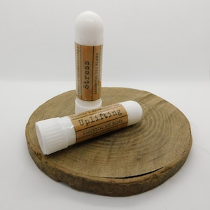 May include: Two white roll-on essential oil blend tubes on a wooden surface. The tubes are labelled "Uplifting Essential Oil Blend" and "Stress Essential Oil Blend".