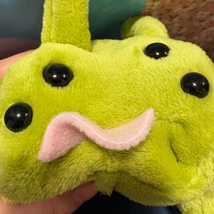 May include: A lime green plush toy with large black eyes and a pink tongue. The toy has a soft, fuzzy texture and a whimsical design, resembling a cartoon character. The toy is held in a hand.
