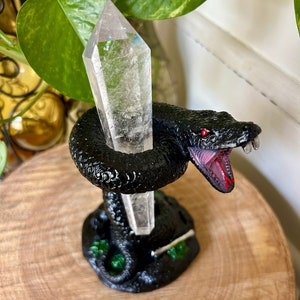 May include: A black snake figurine with a clear quartz crystal point held in its mouth. The snake is standing on a green base and is on a wooden surface.