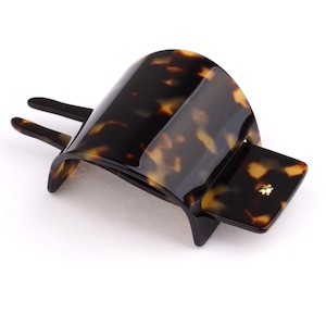 Peut inclure: Une pince à cheveux en écaille de tortue. La pince présente un design incurvé et arqué avec deux broches et un élément carré. Fabriquée dans un matériau brillant et moucheté aux tons bruns et ambrés.