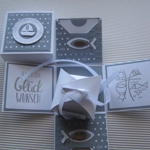 Könnte beinhalten: Eine weiße und graue Papierbox mit einem Pop-up-Design. Die Box ist mit einem Fisch, einem Boot und dem Text "Gott segne dich" und "Glückwunsch" verziert.
