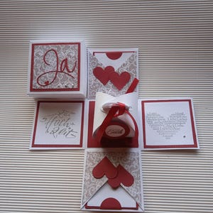 Könnte beinhalten: Eine handgefertigte, offene, weiße und rote Explosionsbox mit Spitze und Herzdetails. Die Box zeigt das Wort "Ja" in roter Schrift und die Worte "Hoch Zeit" in silberner Schrift. Ein rotes Band ist um ein zentrales Element gebunden.