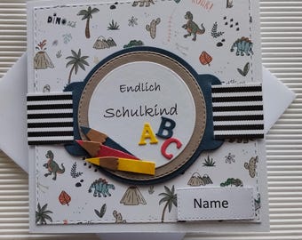 May include: A handmade greeting card with a dinosaur theme. The card features a patterned background with dinosaurs, palm trees, and volcanoes. It includes the German text "Endlich Schulkind" and colorful pencils and ABC letters.