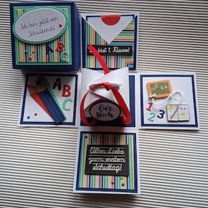 Könnte beinhalten: Eine handgefertigte, mehrschichtige Papierbastelbox mit Schulthema. Die Box ist mit buntem Papier, Bändern und deutschem Text verziert, darunter "Ich bin jetzt ein Schulkind!" und "Alles Liebe zum ersten Schultag!" Die Box enthält Miniatur-Schulelemente.