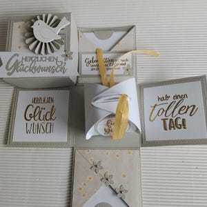 Könnte beinhalten: Eine handgefertigte beige-weiße Explosionsbox mit einer zentralen weißen Kissenverpackung, die mit einem gelben Band verschlossen ist. Die Seiten zeigen deutschen Text wie "Herzlichen Glückwunsch" und "hab einen tollen TAG!" sowie Papierblumen und Vogelmotive.