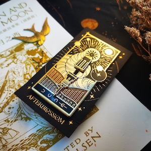 May include: Gold-colored enamel pin with a celestial design, featuring a sun, moon, and stars, on a black card. The pin has the word "JUNE" and "AFTERBLOSSOM" on it. A gold bird-shaped pin and a book with Chinese and English text are also visible.