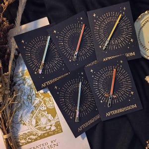 Puede incluir: Una colección de pines de esmalte con diseños de sables de luz en varios colores, incluyendo rojo, amarillo y azul. Cada pin se muestra en una tarjeta negra con un diseño dorado de estrella y el texto "AFTERBLOSSOM".