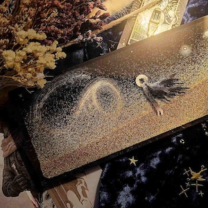 Pode incluir: Uma obra de arte retangular com tema celestial. Apresenta um fundo preto e dourado com uma figura alada e padrões giratórios. A obra de arte é embelezada com glitter dourado e estrelas.