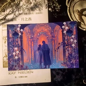 May include: A postcard featuring an illustration of a couple standing in an archway with an orange and blue background. The postcard is on top of a book with text in English and Japanese. The artist's name, Kay Nielsen, is visible.