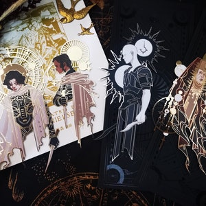 May include: Artistic tarot card designs featuring detailed illustrations of figures with celestial and gothic elements. The cards showcase a color palette of gold, black, and white, with intricate line work and stylized imagery. The cards have a fantasy theme.