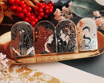 May include: Four decorative metal pins with arched tops, each featuring a unique design. The pins are displayed on a gold-colored tray. The designs include figures, botanical elements, and a snake, with a color palette of black, red, and gold.
