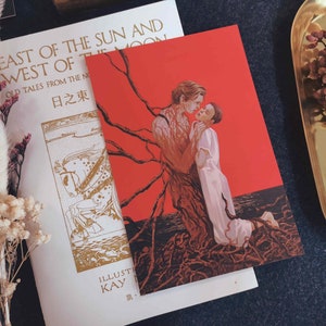 May include: A red art print depicting two figures embracing, with a tree-like design. Beside it is a book titled "East of the Sun and West of the Moon" with gold lettering. Dried flowers and a gold tray are also present.