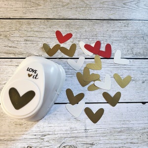May include: A white heart-shaped paper punch with the words "LOVE it" on the side. The punch is surrounded by a variety of heart-shaped paper cutouts in gold, white, and red.