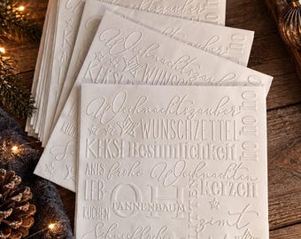 Könnte beinhalten: Ein Stapel quadratischer, weißer Papierservietten mit geprägten weihnachtlichen Wörtern und Sternsymbolen. Der Text enthält Begriffe wie Weihnachtzauber, Wunschzettel, Kekse, Besinnlichkeit, Anis, frohe Weihnachten, Lebkuchen, Tannenbaum und Schneeflocken.