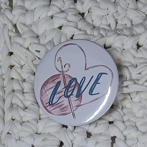 May include: A white button featuring a pink yarn ball and knitting needles, a heart shape, and the word "LOVE" in blue lettering.