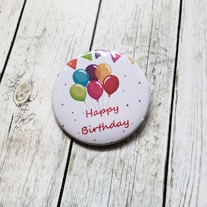 Könnte beinhalten: Runder weißer Knopf mit einer bunten Illustration von Ballons und dem Text "Happy Birthday".