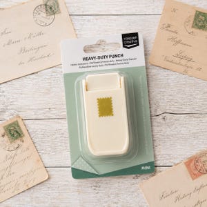 Könnte beinhalten: Ein cremefarbener Heavy-Duty-Stanzer von Vaessen Creative in der Originalverpackung. Das Werkzeug hat einen rechteckigen Körper mit einem briefmarkenförmigen Symbol auf der Vorderseite. Die Verpackung ist weiß und salbeigrün mit dem Text HEAVY-DUTY PUNCH.