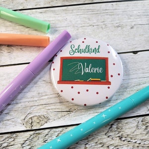 Könnte beinhalten: Ein weißer Knopf mit roten Punkten und dem Text "Schulkind Valerie" in Grün und Weiß. Der Knopf hat ein Tafeldesign. Pastellfarbene Marker in Grün, Orange, Lila und Türkis sind um den Knopf herum verstreut.