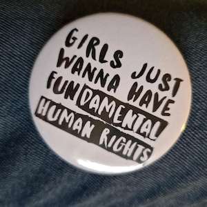 Könnte beinhalten: Ein runder weißer Ansteckbutton mit schwarzem Text, der GIRLS JUST WANNA HAVE FUNDAMENTAL HUMAN RIGHTS lautet. Das Wort FUNDAMENTAL ist mit einem schwarzen rechteckigen Hintergrund hervorgehoben und der Text ist in einem fetten, handgeschriebenen Stil.
