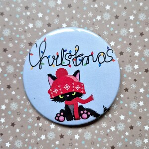 Könnte beinhalten: Ein runder, hellblauer Button mit einer schwarzen Katze, die einen roten Hut und Schal trägt. Über der Katze steht das Wort "Christmas" mit bunten Lichtern. Der Hintergrund ist ein Muster aus Schneeflocken und Sternen.
