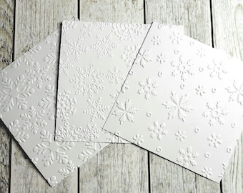May include: Four white embossed cards with snowflake designs. The cards are arranged in a fan shape on a light wood background.