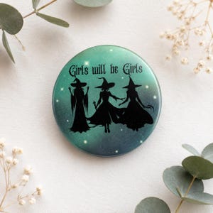 Könnte beinhalten: Ein runder Ansteckbutton mit einem Farbverlauf von Türkis zu Dunkelblau und einem Sternenhimmel-Effekt. Das Design zeigt drei schwarze Silhouetten von Hexen mit spitzen Hüten und langen Kleidern sowie den Schriftzug Girls will be Girls.