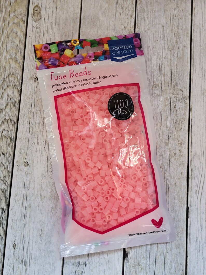 May include: A bag of 1100 pink fuse beads, also known as ironing beads, for crafting. The bag is labeled "Fuse Beads" and has the brand name "vaessen creative" printed on it.