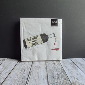 Könnte beinhalten: Ein Stapel weißer Papierservietten mit einer Weinflaschenillustration. Die Flasche gießt rote Flüssigkeit aus, mit dem deutschen Text "DER KLUGERE kippt NACH". Die Servietten haben das "ppd"-Logo oben rechts.