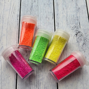 Könnte beinhalten: Sechs kleine, durchsichtige Plastikbehälter gefüllt mit buntem Glitter. Die Behälter sind in einem Halbkreis auf einer hellen Holzoberfläche angeordnet. Die Glitterfarben sind rosa, rot, gelb, grün und orange.