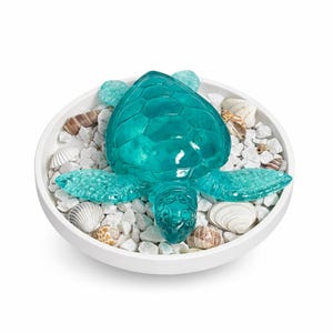 Könnte beinhalten: Eine türkisfarbene Glas-Schildkrötenfigur mit detailliertem Panzer und Flossen, in einer weißen Schale mit weißen Kieselsteinen und Muscheln. Dekorative Figur für Küsten- oder Stranddekoration.