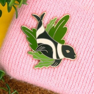 May include: A gold-colored enamel pin featuring a black and white fish with green seaweed. The fish has a white stripe down its side and a black eye.